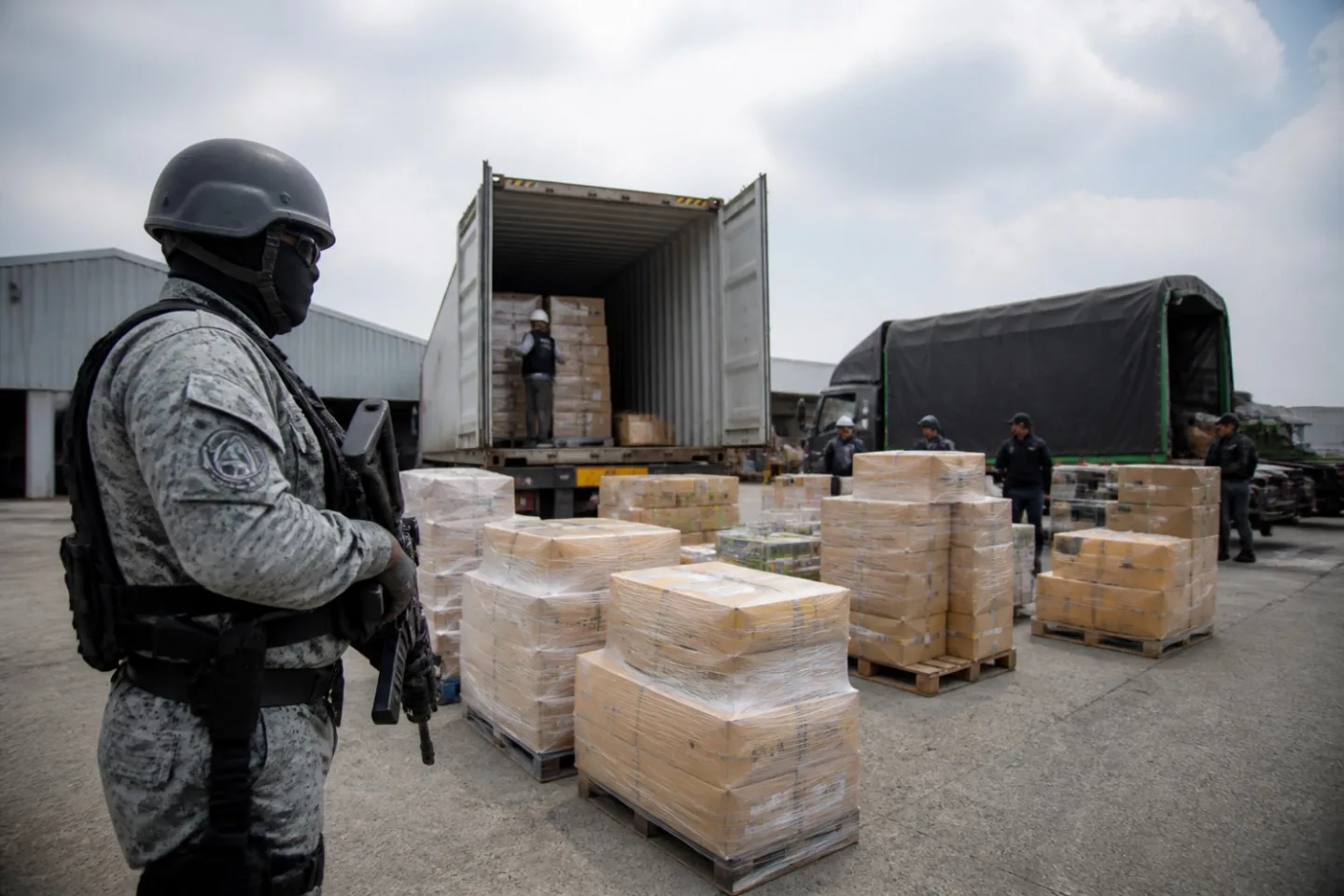 Una vista amplia de un almacén grande muestra a agentes de la DIAN revisando pilas de cajas que contienen ropa y zapatos. La escena está bien iluminada por una luz natural difusa que entra por las ventanas superiores, creando un ambiente de inspección minuciosa.