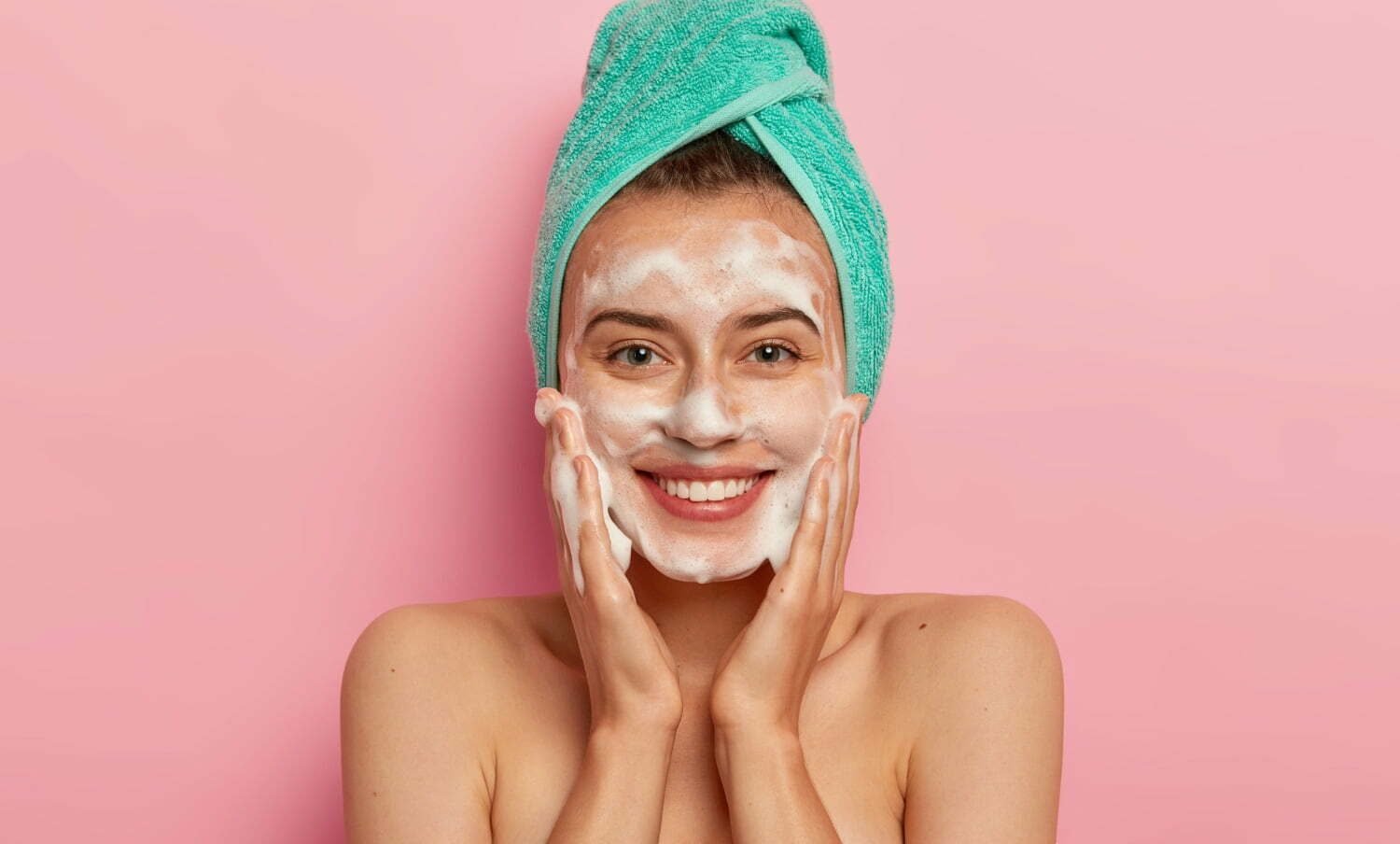 mujer sonriente con toalla en la cabeza y jabón en la cara, con las manos en sus mejillas - lavarte la cara en la ducha