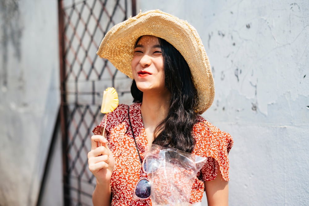 Joven mujer viajera tailandesa hermosa vestida de rojo y que disfruta comiendo pedazos de piña con un palo / piña